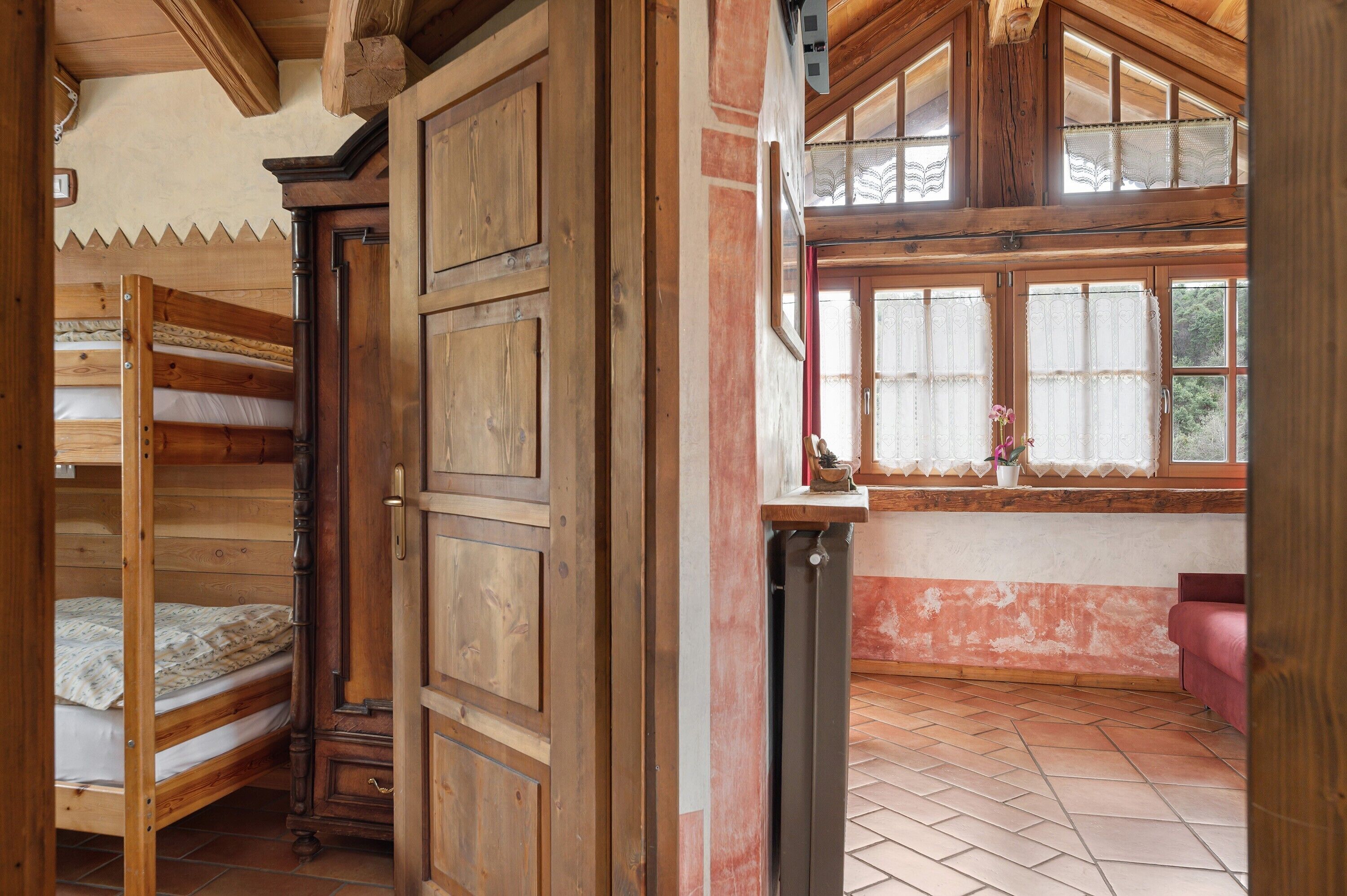 Une pièce avec des lits superposés en bois, un radiateur, une fenêtre ornée de rideaux en dentelle et un canapé rouge.
