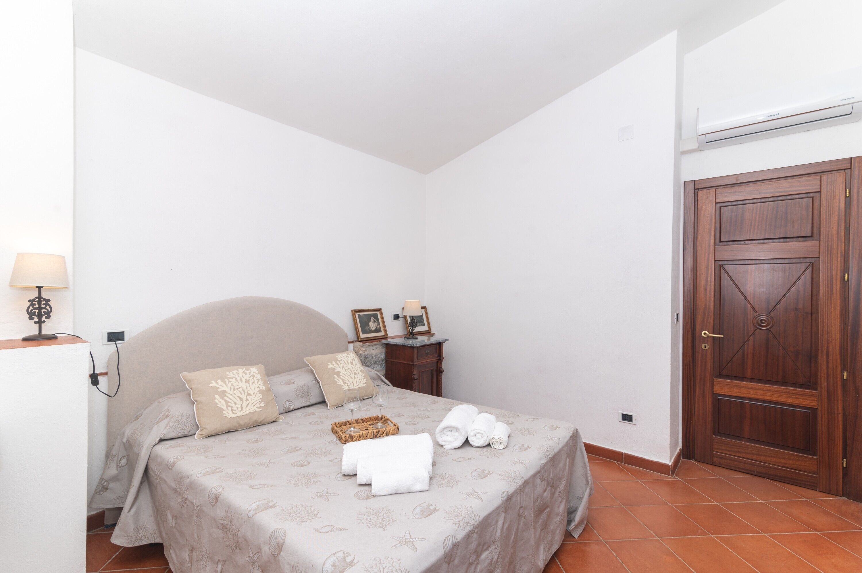 A neatly made bed with towels, a bedside lamp, and a small table with a lamp and picture frames.