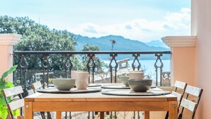 A wooden table with a view of the sea and mountains, a balcony with a railing, and a potted plant.