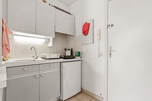 A compact kitchen with white cabinets, a sink, a kettle, and a red oven mitt on the wall.