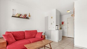 Un canapé rouge, une table en bois, un espace cuisine blanc et un mur blanc avec une étagère.