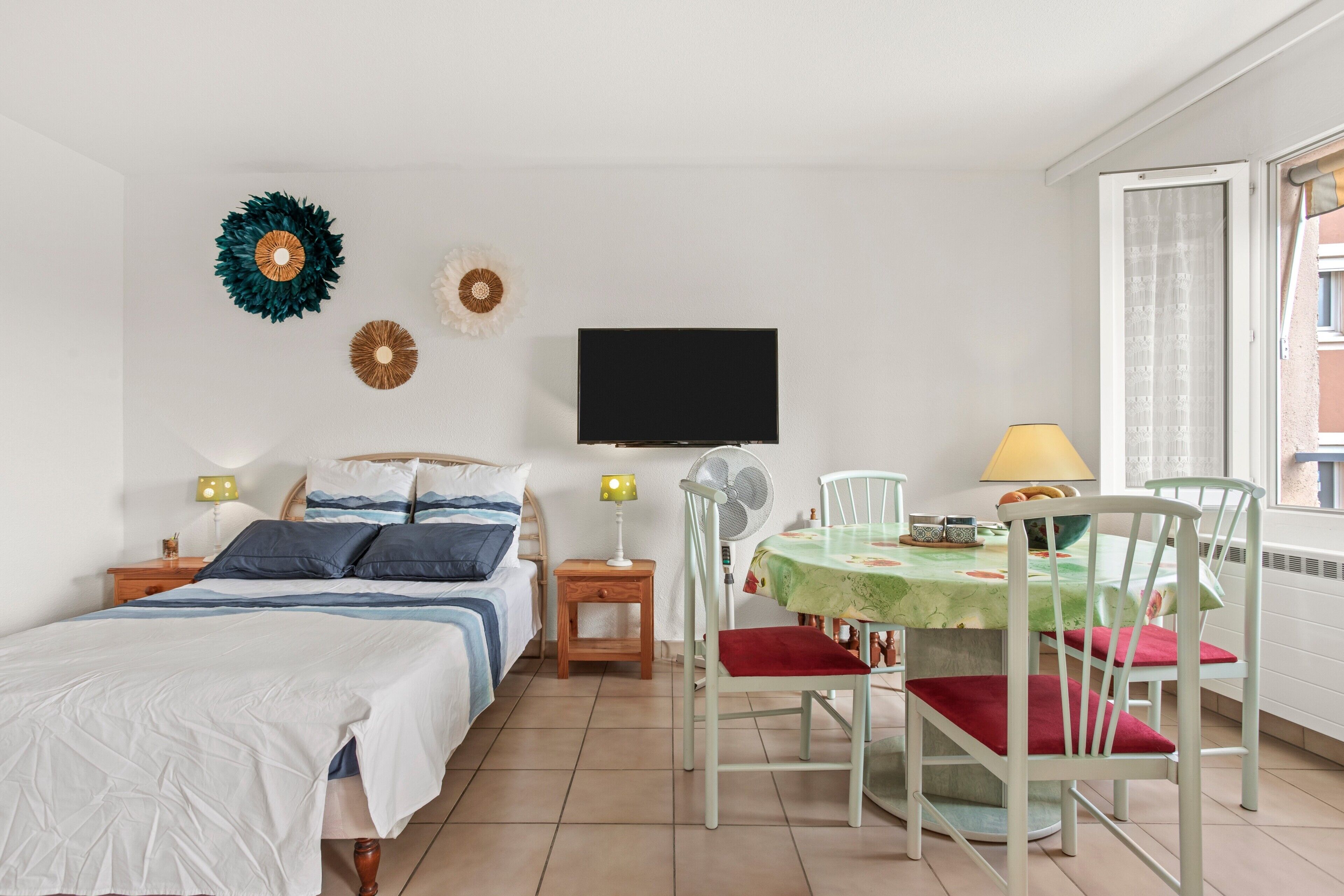 A bedroom with a bed, bedside tables, a TV, a fan, and a dining area with chairs and a table.
