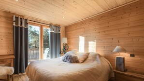 A wooden-paneled bedroom with a large bed, a nightstand, a lamp, and a view through a sliding door. - Chalet 'Velajule', private shuttle at the slopes, private jacuzzi & shared pool (Manigod)