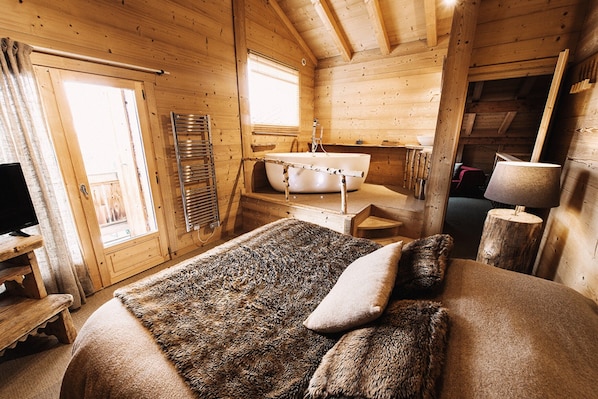 A wooden cabin interior with a large bed, a wooden bench, a bathtub, and a window.