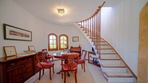 Un área de comedor con una mesa redonda y sillas, una escalera con barandilla de madera y dos ventanales arqueados.