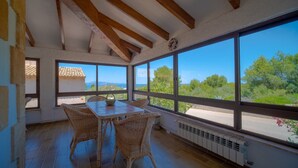 Una habitación con un ventanal grande, vigas de madera, muebles de mimbre y vistas a zonas verdes y edificios.