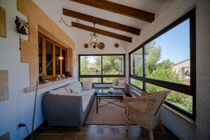 A sunlit room with a sofa, wicker chair, and a table, featuring large windows and a ceiling fan.