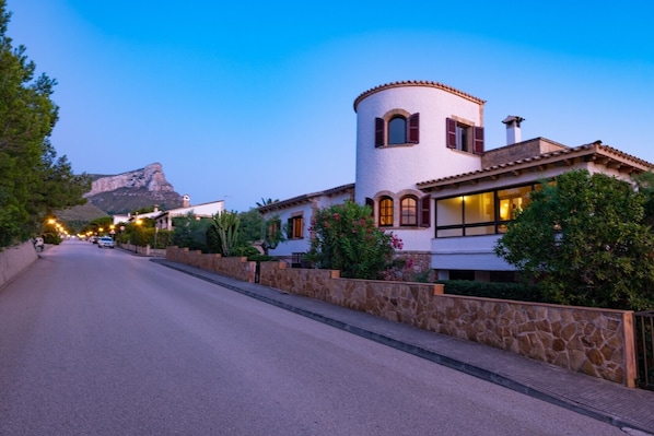 A street with a white building featuring a round tower, stone walls, and a mountain in the background.