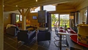 A wooden interior with a fireplace, seating area, dining table, and large windows. - Chalet 'Pesse' with Mountain View, Private Jacuzzi & Shared Pool (Summer Only) (Manigod)