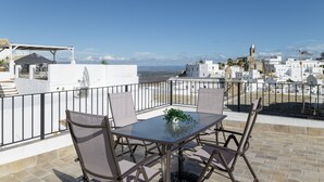 A rooftop terrace with a table and chairs, a potted plant, and a view of a coastal town with white buildings and a church.