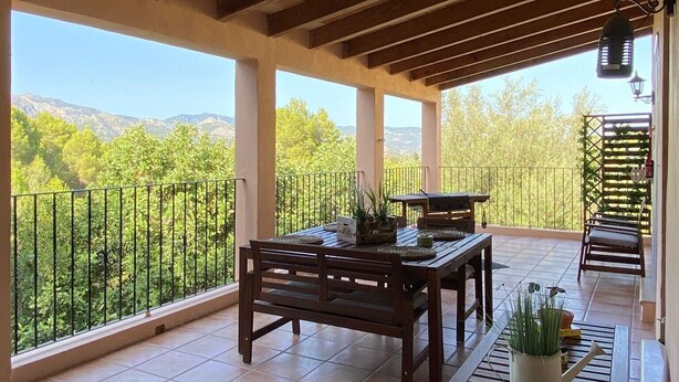 Une terrasse couverte avec une table et des chaises en bois, un pot de fleurs et une vue sur la verdure et les collines.