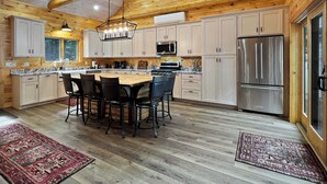 A spacious kitchen with wooden walls, a large island with chairs, stainless steel appliances, and a hanging light fixture. - Sunset Serenity (Waterfront on Moosehead Lake!) (Greenville)