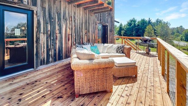 A wooden deck with a wicker sofa and cushions, a grill, and a view of the surrounding forest. - Lake View Lodge (Waterfront in Town!) (Greenville)