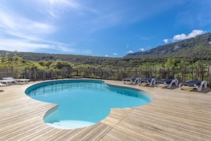 Una terraza de madera con piscina, sillas de descanso y montañas al fondo.