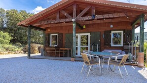 A wooden cabin with a covered patio, outdoor seating, and a gravel ground.