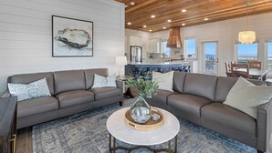 A modern living room with a brown leather sofa, a round coffee table with decorative items, and a wooden ceiling. - NEW HOME! AMISTAD IS AN UPSCALE 6BD/4BA HOME W/GULF & CANAL VIEWS, PRIVATE POOL! (Mexico Beach)