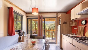 A modern kitchen with a dining area, wooden cabinets, a red clock, and a view of a wooden deck. - Chalet 'Canada' with Mountain Views, Shared Pool and Private Garden (Rouffiac-des-Corbières)