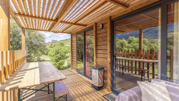 A wooden deck with a table and bench, a trash can, and a sofa with a view of the outdoors. - Chalet 'Canada' with Mountain Views, Shared Pool and Private Garden (Rouffiac-des-Corbières)