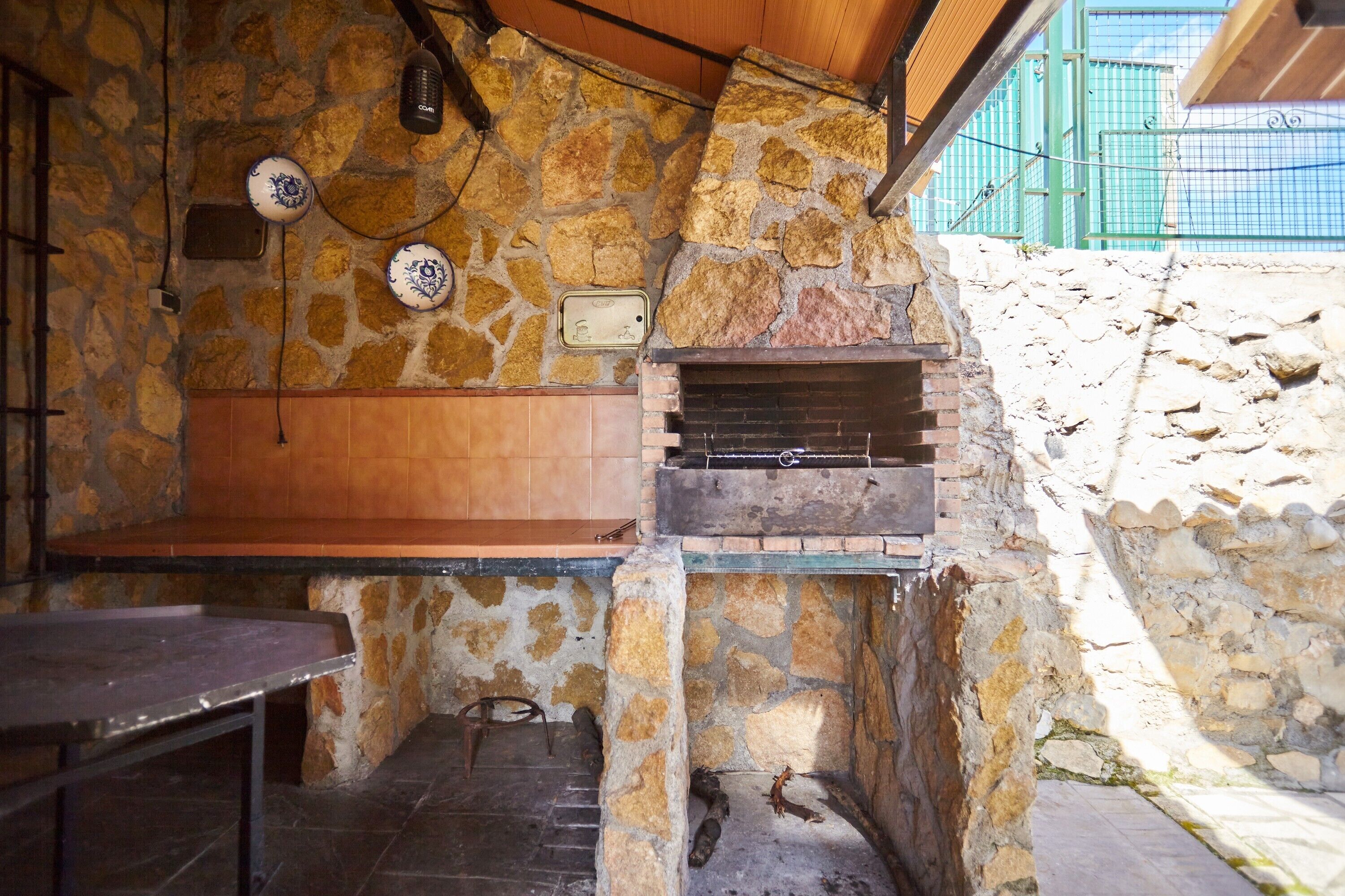 Un área de cocina al aire libre con paredes de piedra, una parrilla empotrada, un banco de madera y una mesa de metal.