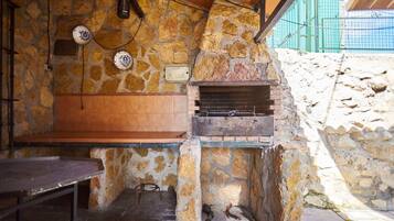 Un área de cocina al aire libre con paredes de piedra, una parrilla empotrada, un banco de madera y una mesa de metal.