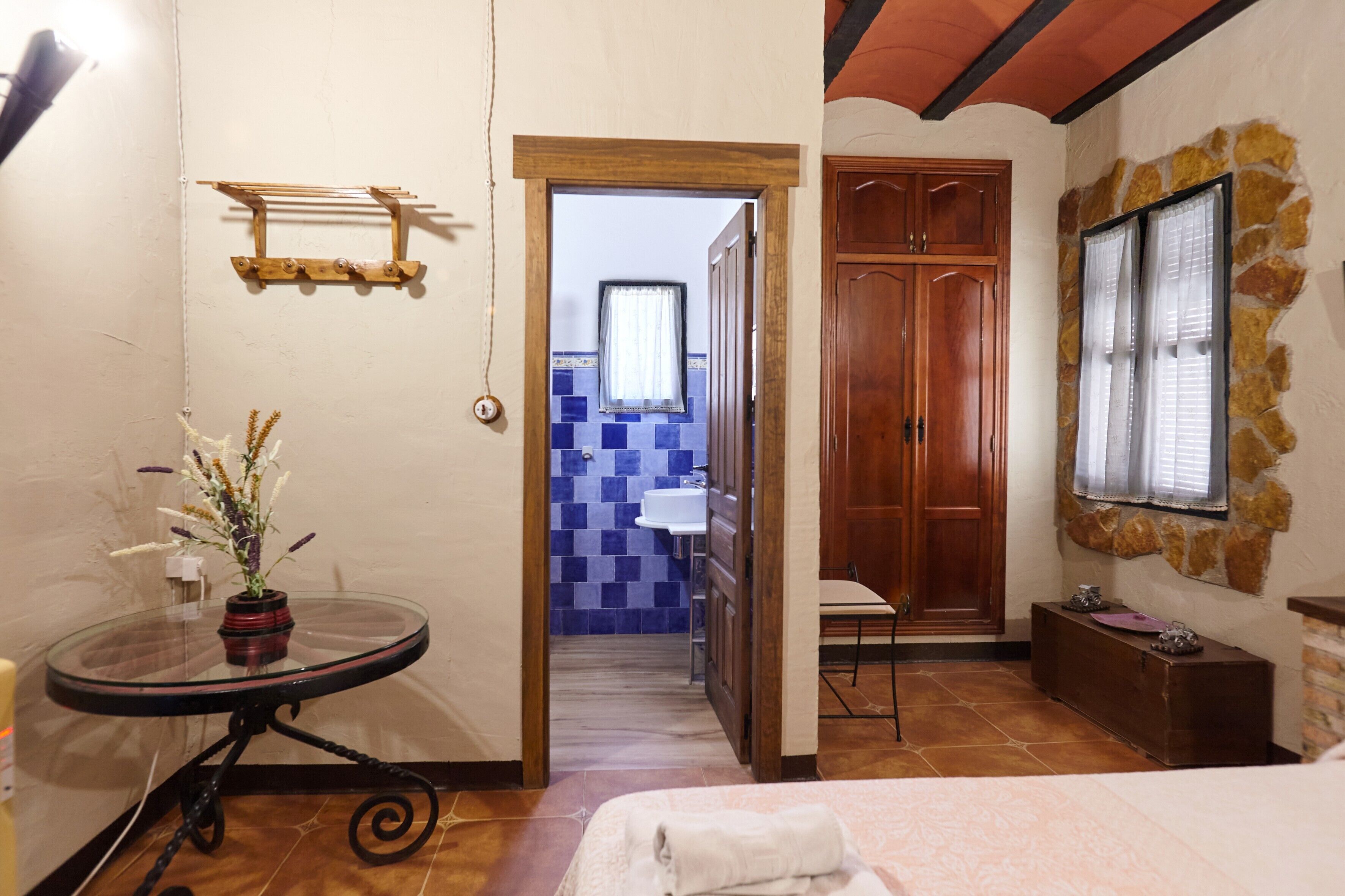 Una habitación con cama, una mesa con un florero, un armario de madera y una pared de piedra.
