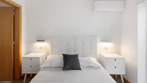 A bedroom with a bed, two bedside tables, a nightstand, a wall-mounted air conditioner, and a door.