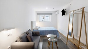 A compact hotel room with a bed, a sofa, a small table, a mirror, and a TV.