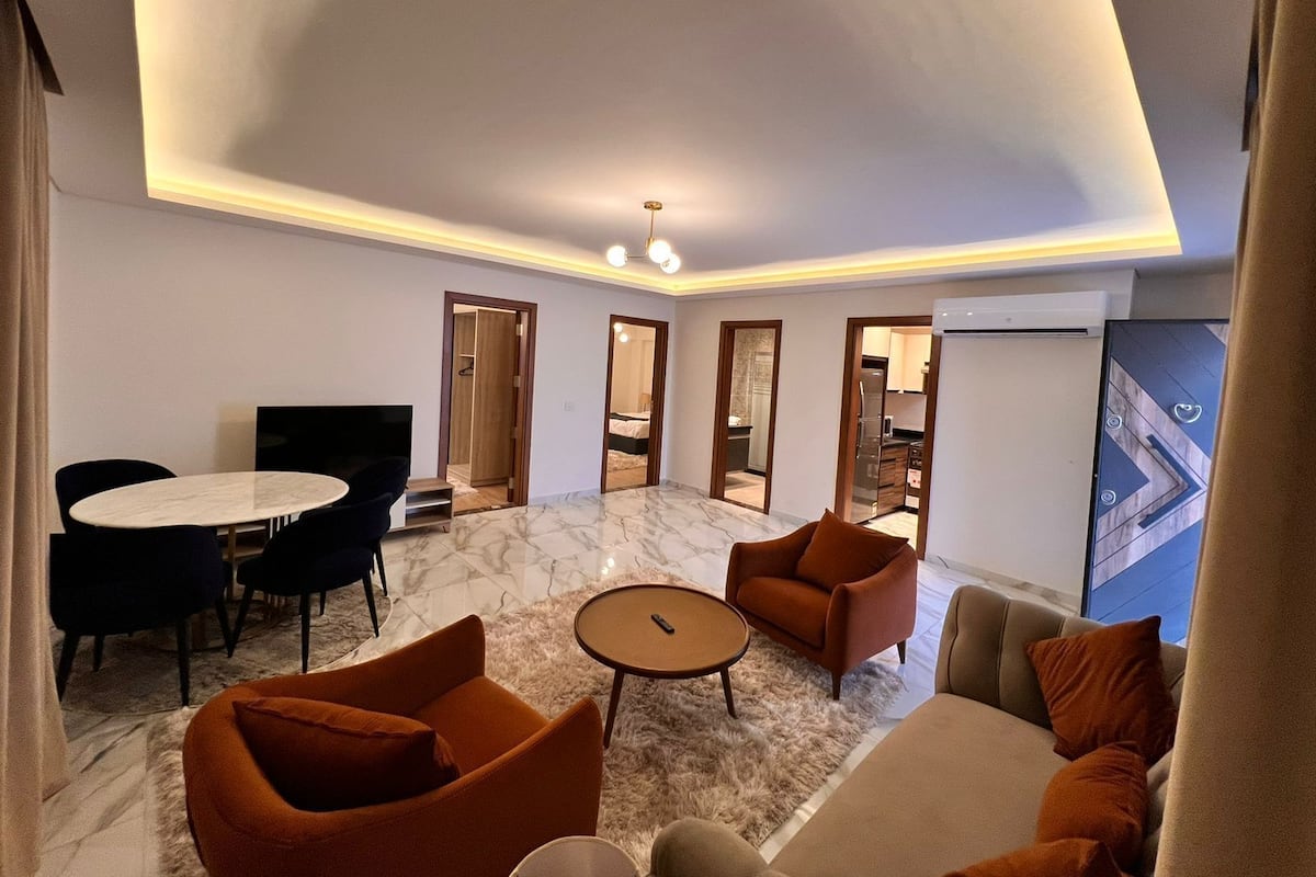 Uma sala de estar moderna com piso de mármore, uma mesa de centro redonda e poltronas laranjas. Há uma mesa de jantar com cadeiras e uma televisão no canto.