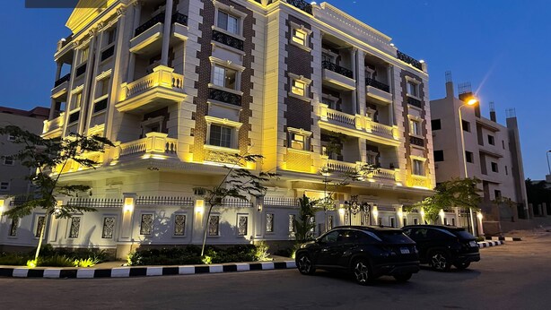 A multi-story hotel with balconies, a well-lit facade, and a parking area with two cars. - White Palace Serviced Apartments (New Cairo)