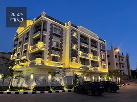 A multi-story hotel with balconies, a well-lit facade, and a parking area with two cars.