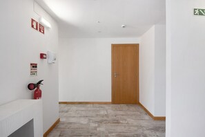 A hallway with a wooden door, white walls, a fire extinguisher, and emergency exit signs. - Room 'Al Katekero - Quarto 104' with Wi-Fi and Air Conditioning (Gafanha da Encarnação)
