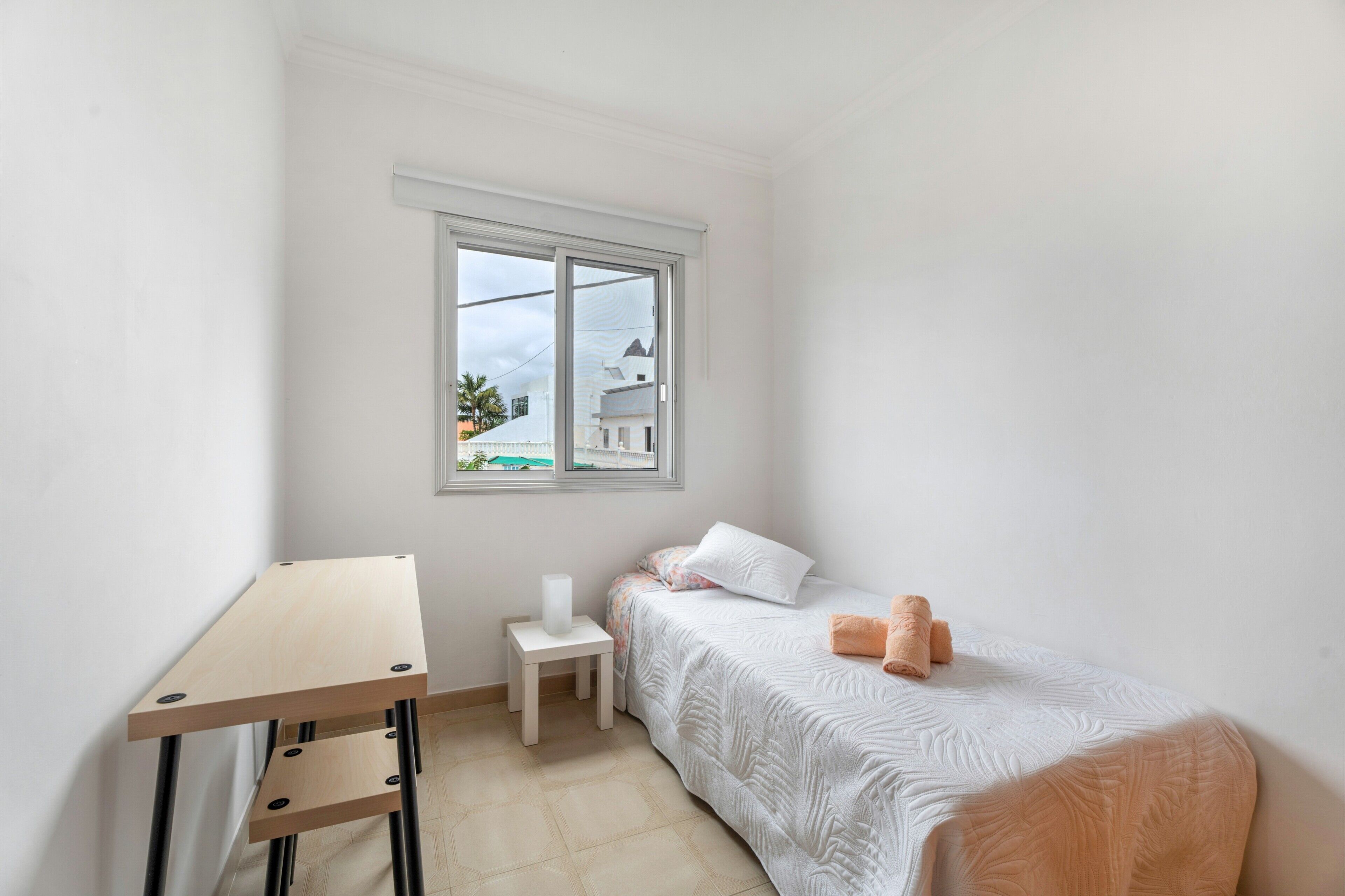 A small, neatly arranged bedroom with a bed, a bedside table, a window, and a desk.
