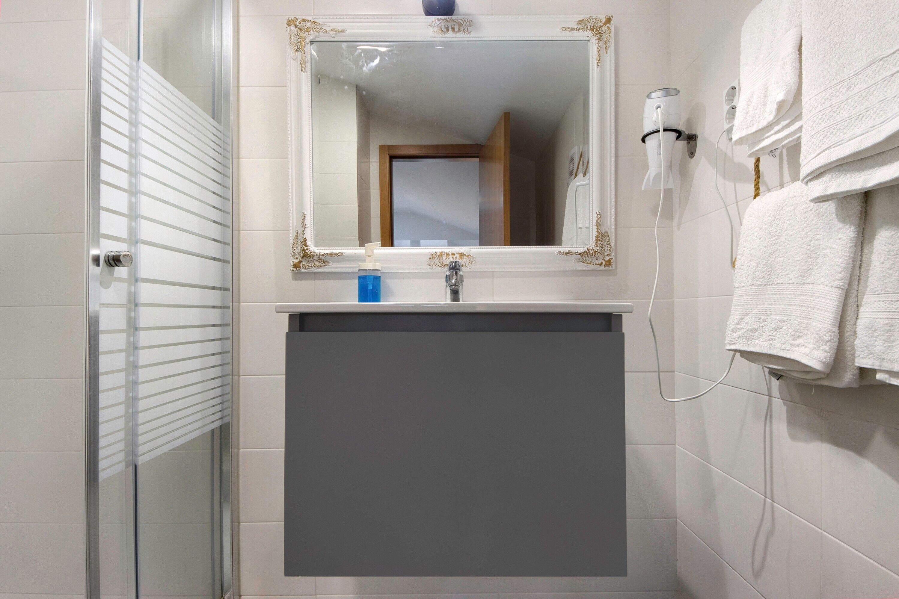 Un baño con cabina de ducha de vidrio, espejo con marco dorado, lavamanos blanco y toallero con toallas dobladas.