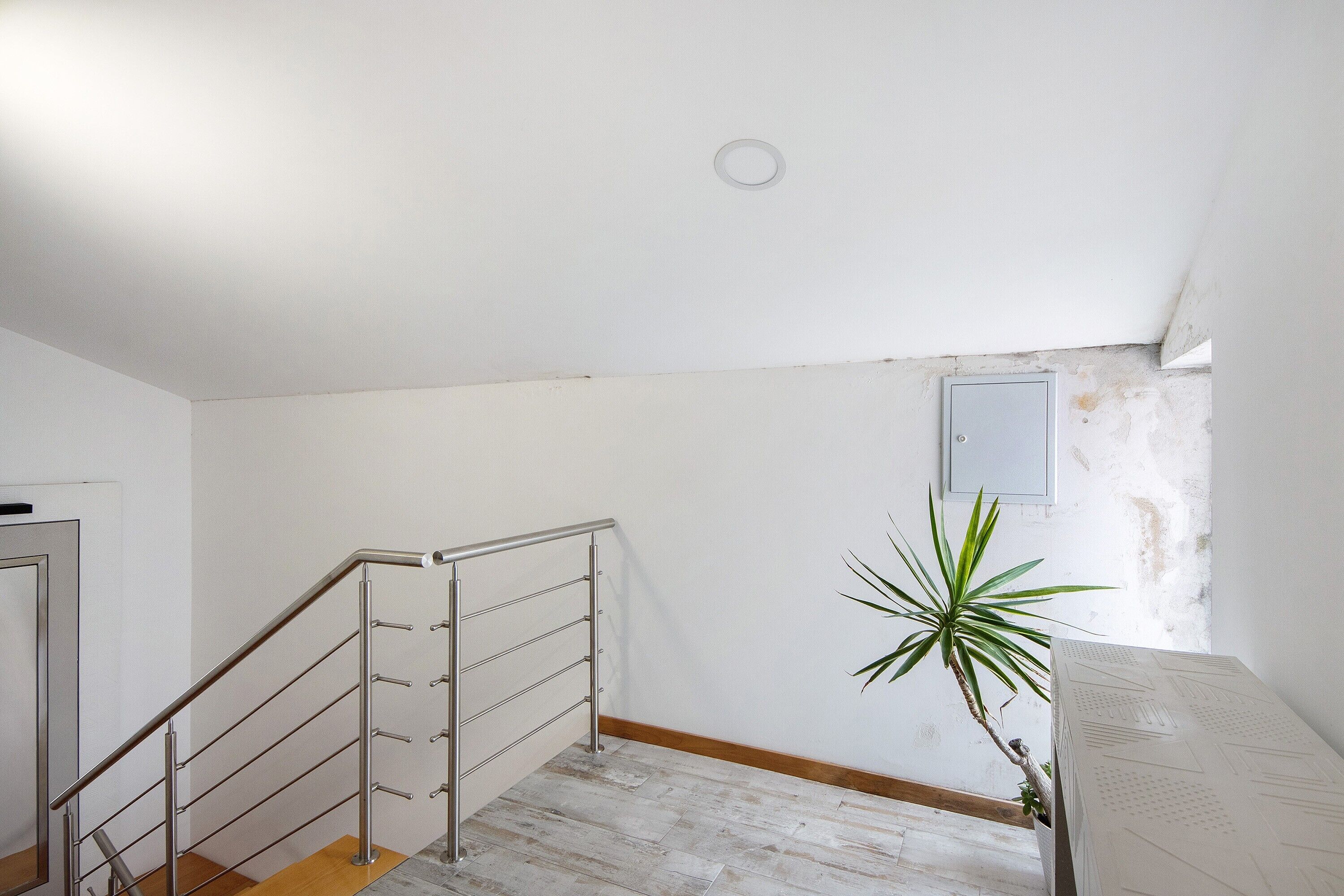 Una escalera con barandas de metal, piso de madera, pared blanca y una planta en maceta.