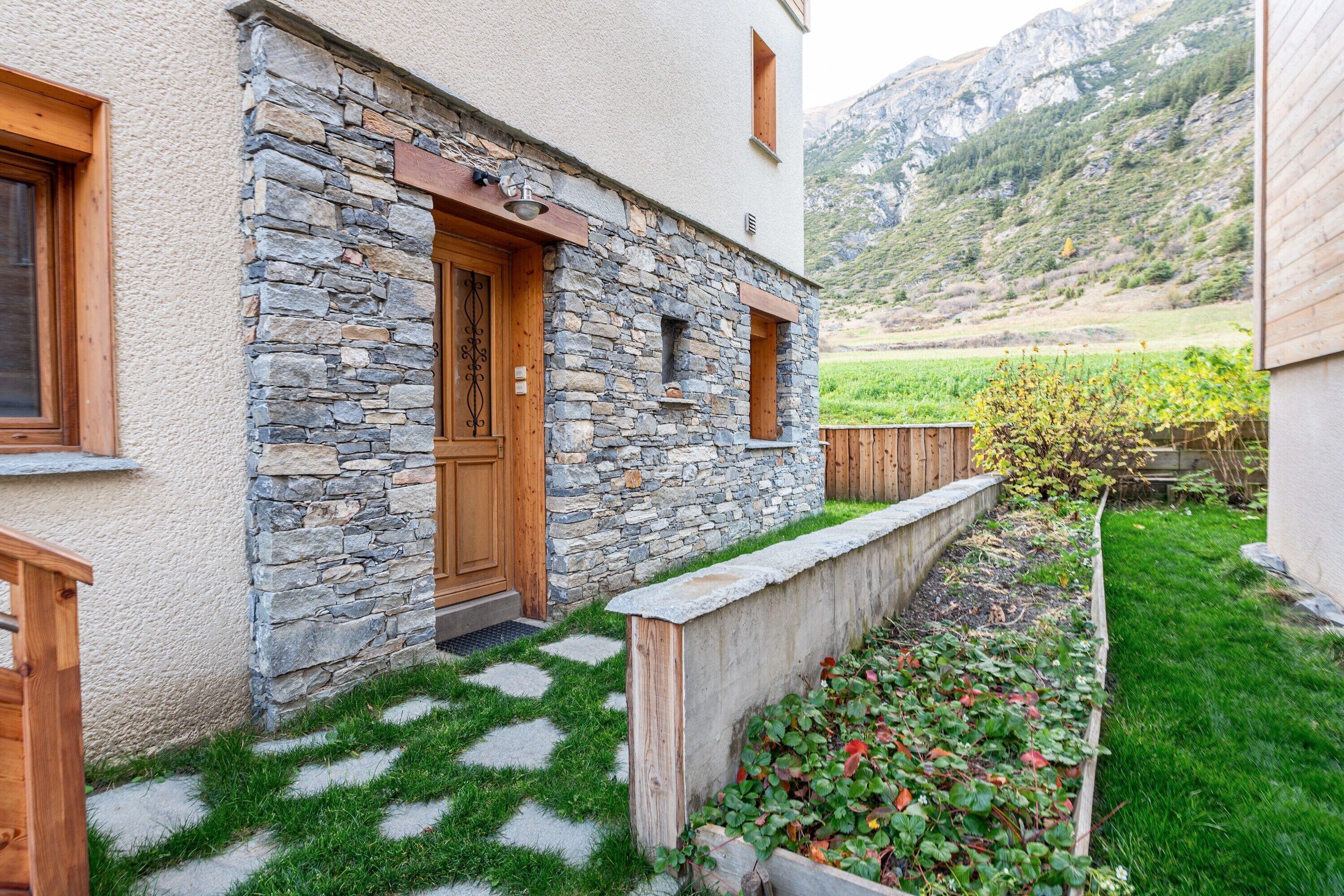 Een stenen gebouw met een houten deur, een klein raam en een tuin met gras en planten.