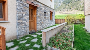Un edificio de piedra con puerta de madera, una ventana pequeña y un jardín con césped y plantas.