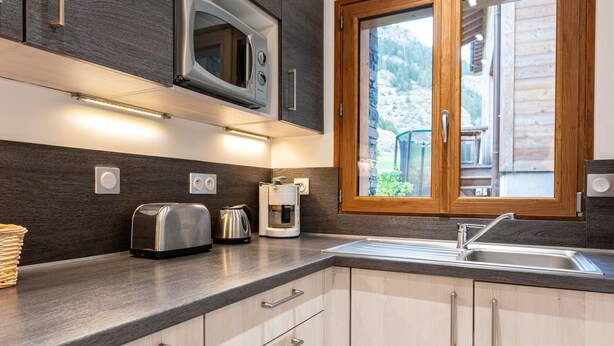 Una cocina moderna con microondas, tostadora y cafetera integrados, un amplio ventanal con vistas a un paisaje montañoso, y un lavabo con grifo de acero inoxidable.