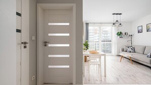 A modern living room with a white sofa, a dining table with chairs, and a large window with a view of buildings. - Warszawa Rakowska Cosy Apartment (Warszawa)