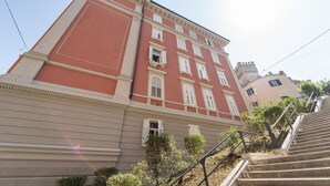 Un edificio de varios pisos con fachada roja, una escalera que lleva a la entrada y un pequeño jardín.