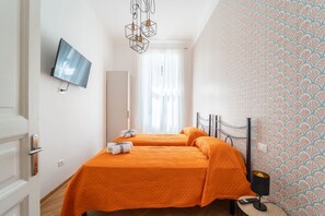 Ein Hotelzimmer mit zwei Betten, einem Fernseher, einem Spiegel, einem Fenster mit Vorhängen, einer Wand mit Muster und einem Nachttisch mit Lampe.