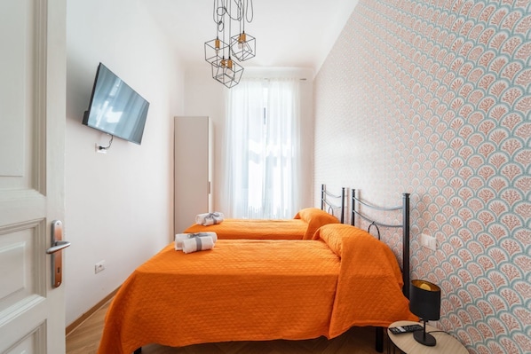 Ein Hotelzimmer mit zwei Betten, einem Fernseher, einem Spiegel, einem Fenster mit Vorhängen, einer Wand mit Muster und einem Nachttisch mit Lampe.
