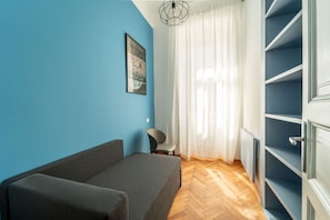 Ein Zimmer mit blauer Wand, einer Couch, einem Sessel, einem Fenster mit durchsichtigen Vorhängen und einer eingebauten Regaleinrichtung.