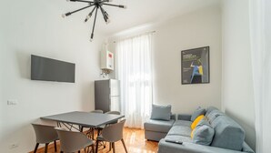 Una sala de estar moderna con un sofá gris, una mesa de comedor con sillas, un televisor de pantalla plana y un candelabro.