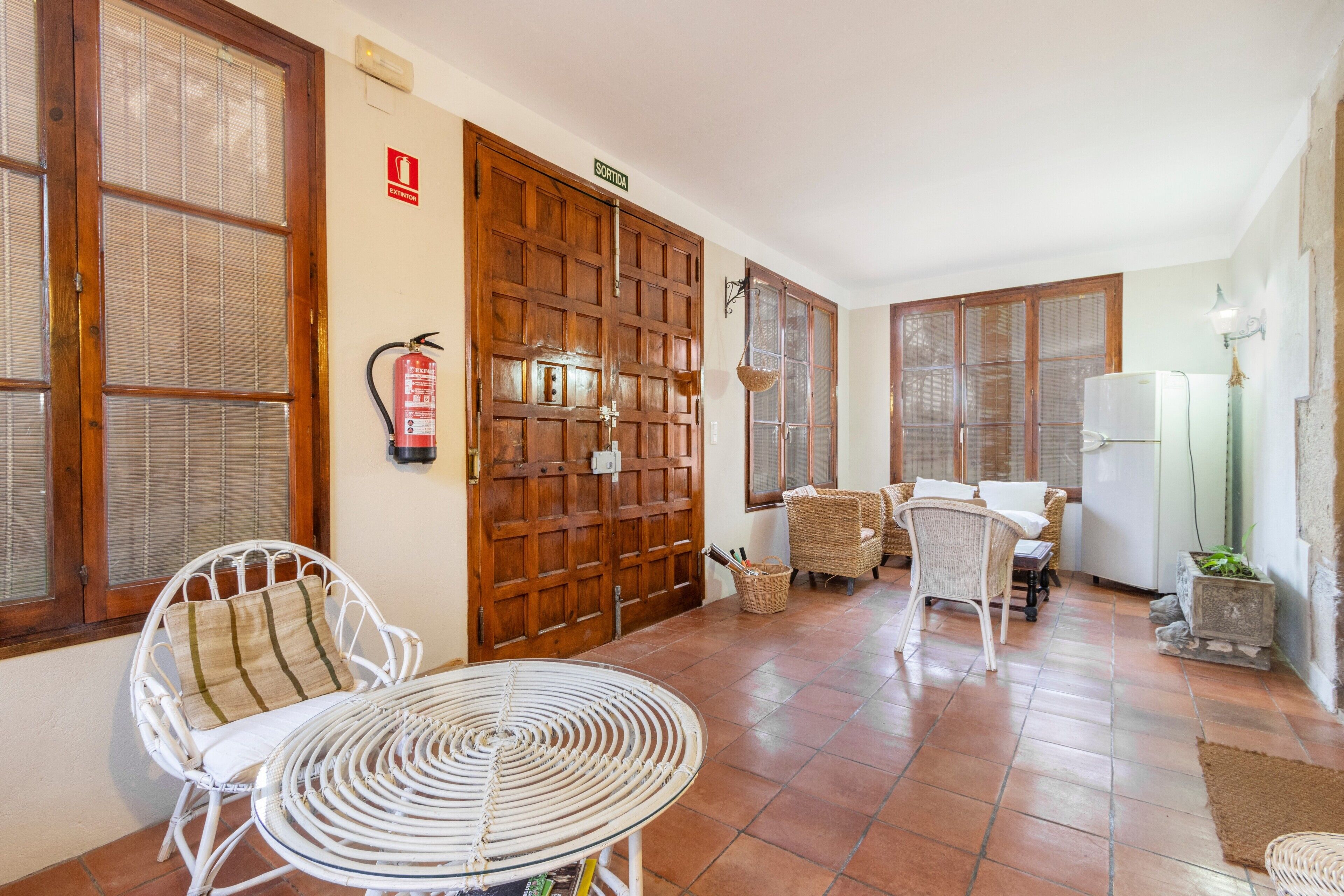 Una habitación con puerta de madera, muebles de mimbre, una mesa blanca y un refrigerador blanco.