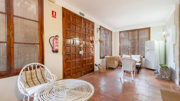 Una habitación con puerta de madera, muebles de mimbre, una mesa blanca y un refrigerador blanco.