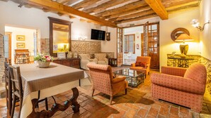 Un intérieur rustique avec des poutres apparentes, une cheminée en pierre, un coin repas avec une nappe à carreaux et des fauteuils à motifs.