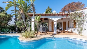 Une piscine bordée de carreaux, un patio couvert avec des chaises et un bâtiment blanc surmonté d’un toit en tuiles.