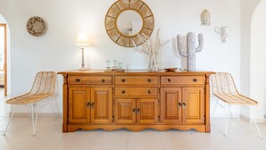 Un meuble en bois doté de tiroirs et de portes, flanqué de deux chaises en osier, d’un miroir rond et d’objets décoratifs accrochés au mur.
