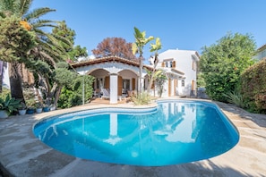 Una piscina con bordeado de azulejos, rodeada por una zona de patio con sillas y una mesa, y un edificio con techo y columnas de azulejos.