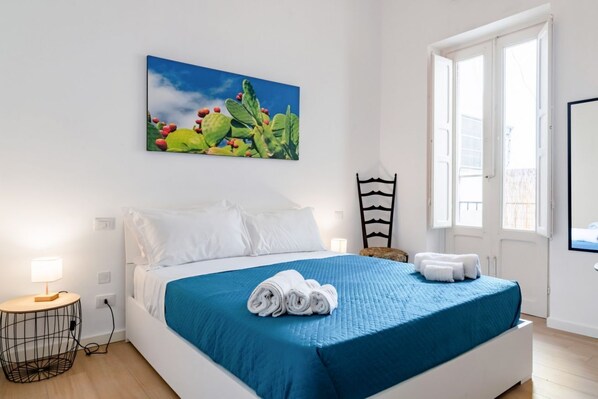 Un dormitorio con cama, almohadas, una colcha azul, una toalla enrollada, una mesita de noche, una lámpara, una silla y una ventana.
